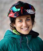 Headshot of Meg Whicher wearing a green zipper jacket and multi-colored bicycle helmet.